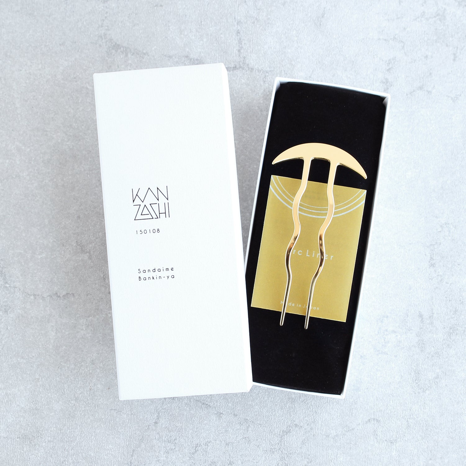 white kanzashi box packaging with a black interior inside a gold kanzashi hairpin on a light gray background.