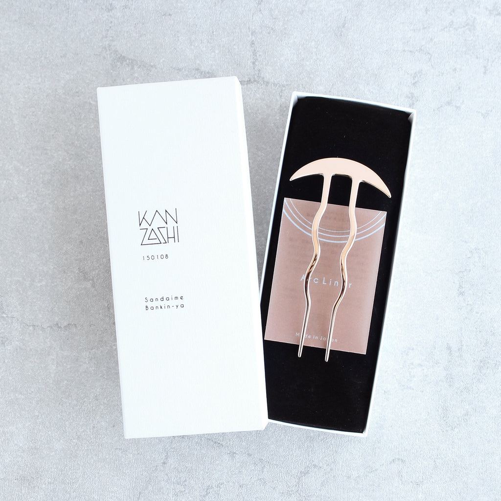 Rose gold kanzashiin the packaching box with white exterior and black interior on a gray background.